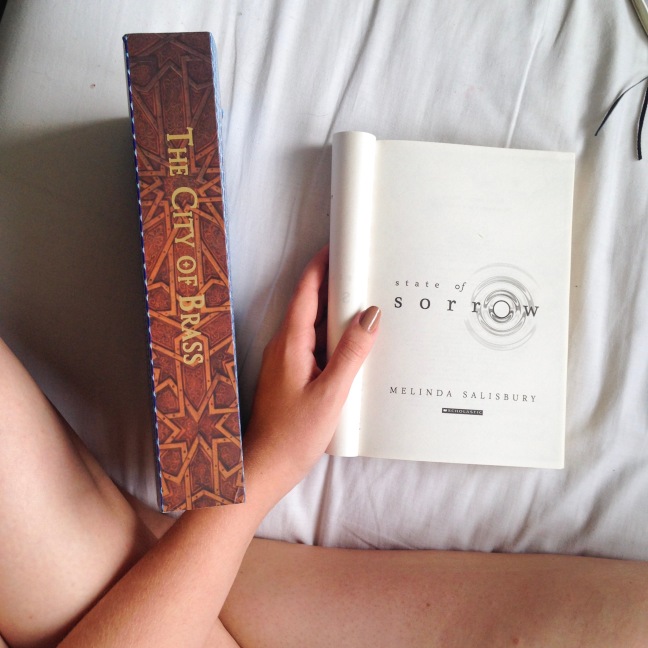 Crossed legs with my hand holding open State of Sorrow by Melinda Salisbury, and The City of Brass by S.A. Chakraborty stood beside it, spine showing